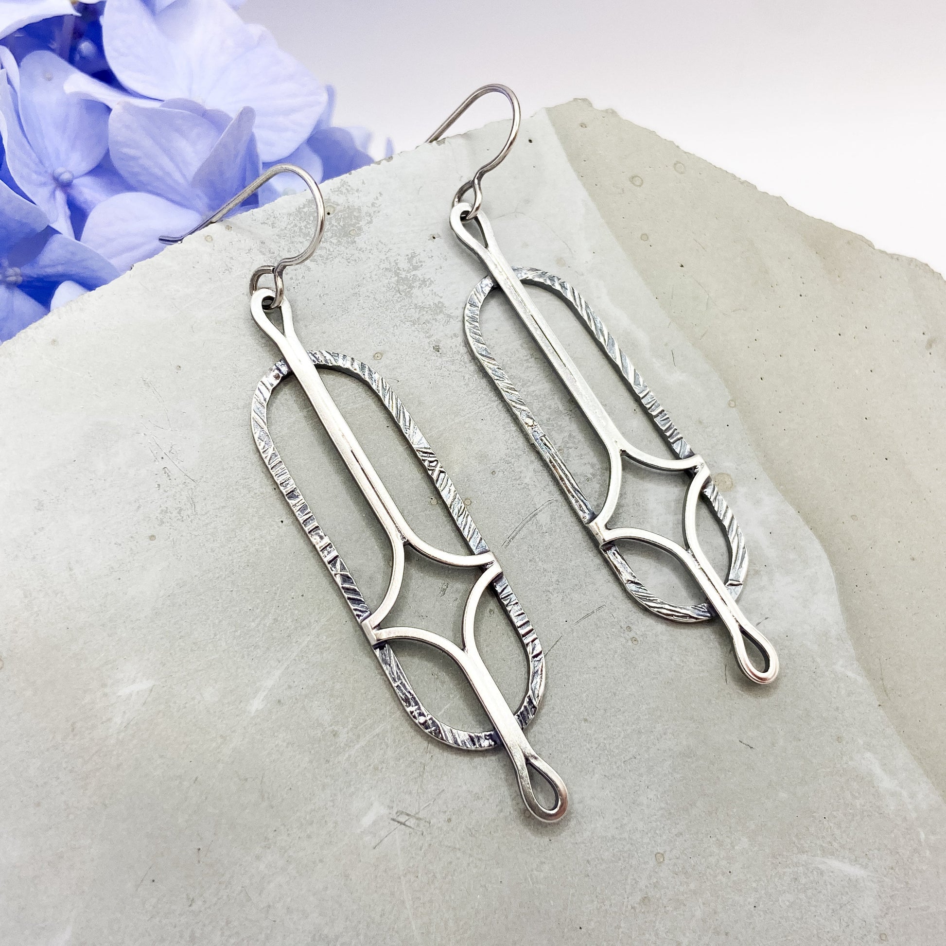 Silver earrings on a stone surface with purple flowers in the background